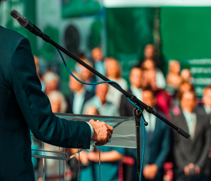 imagen que muestra como una especia de speech, hay varias personas al frente, se muestra al costado izquierdo una persona posando sus manos en un atril de vidrio, va bien vestido con saco y tiene un micrófono a su lado derecho.